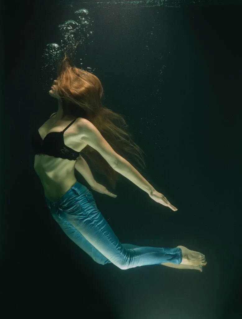 a red haired woman floating underwater in jeans and a black bra.