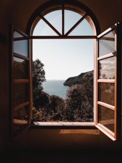 an old wooden window open to a view of the ocean and trees.