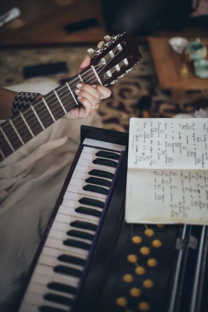 woman's hand on the neck of a guitar next to a piano and sheet music