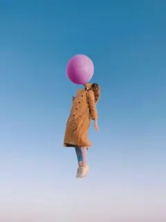 girl in a trenchcoat floating in the sky while blowing large pink bubble gum bubble.