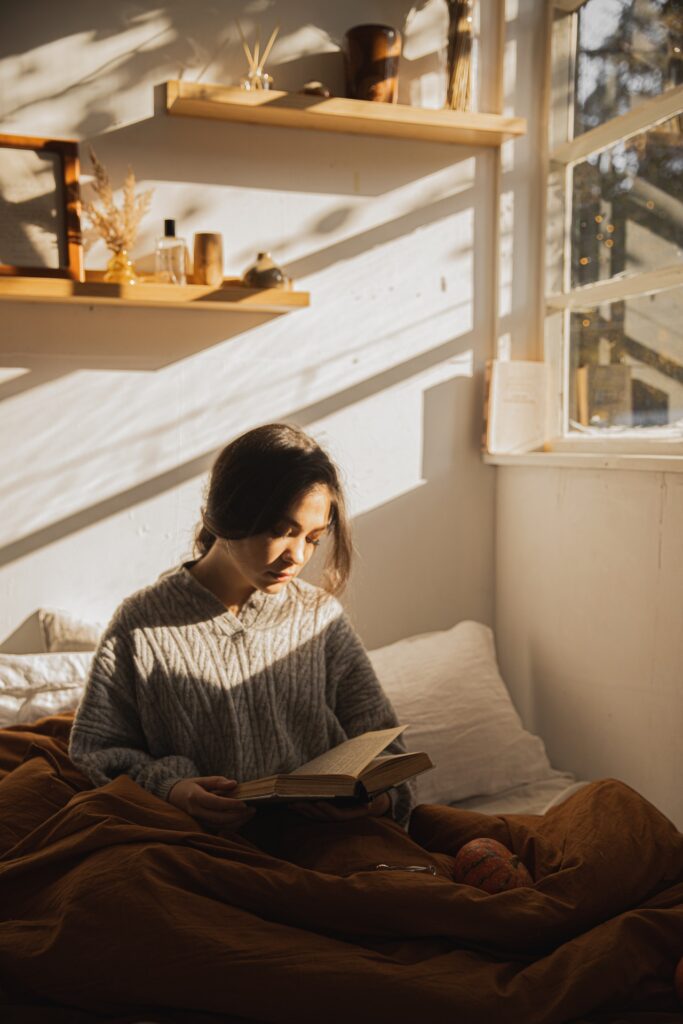 young brunette woman in a gray sweater reading a book in bed.