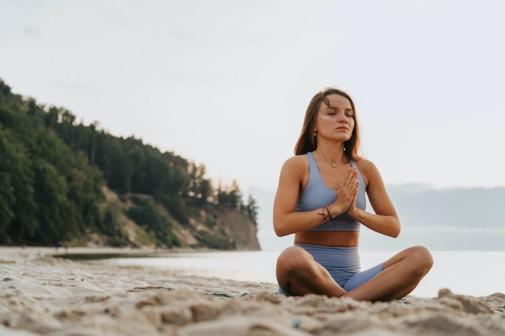 young brunette woman in gray workout attire meditating on a beach with pine trees in the background.